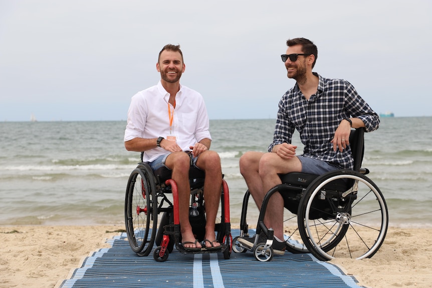 Two men sit side by side in wheelchairs, on a beach. 