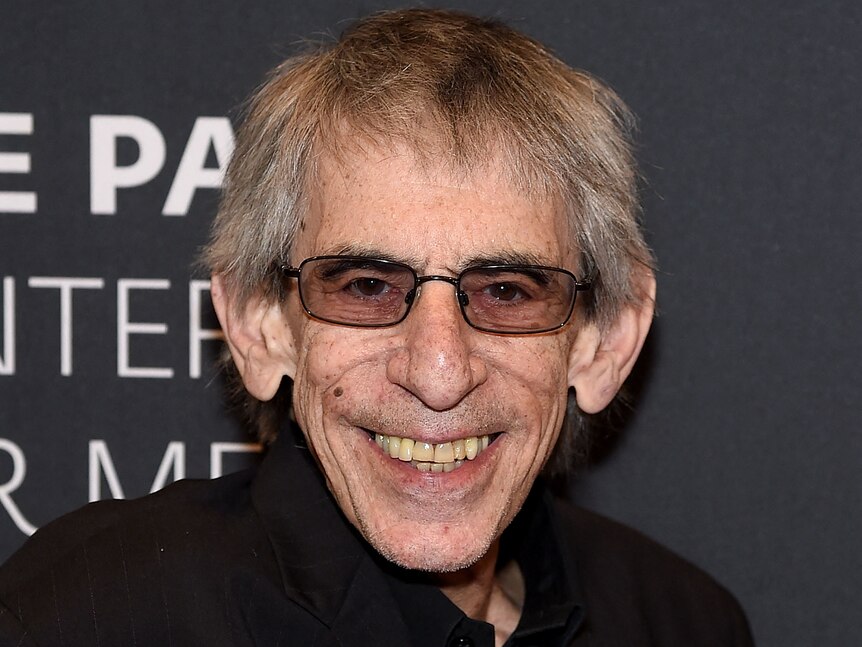 Richard Belzer smiles wearing glasses in front of a black background. 