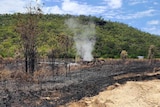 Burnt out land in front of hill with dark patches, smoke rising, blue sky with few clouds.