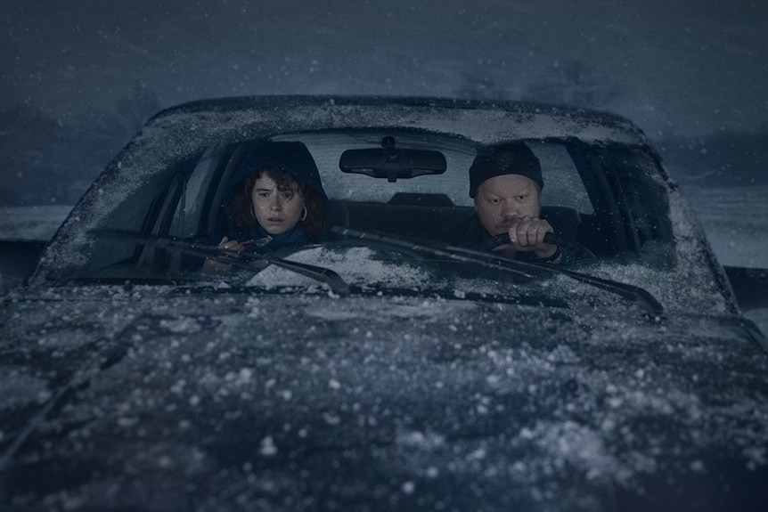A young worried looking woman sits with man in car covered in snow as a blizzard rages on behind them in a dark gloomy sky.