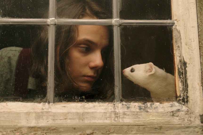 A girl stares at a white ferret as they sit by a window.