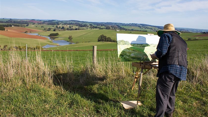 A man painting at an easel, looking out over a valley landscape