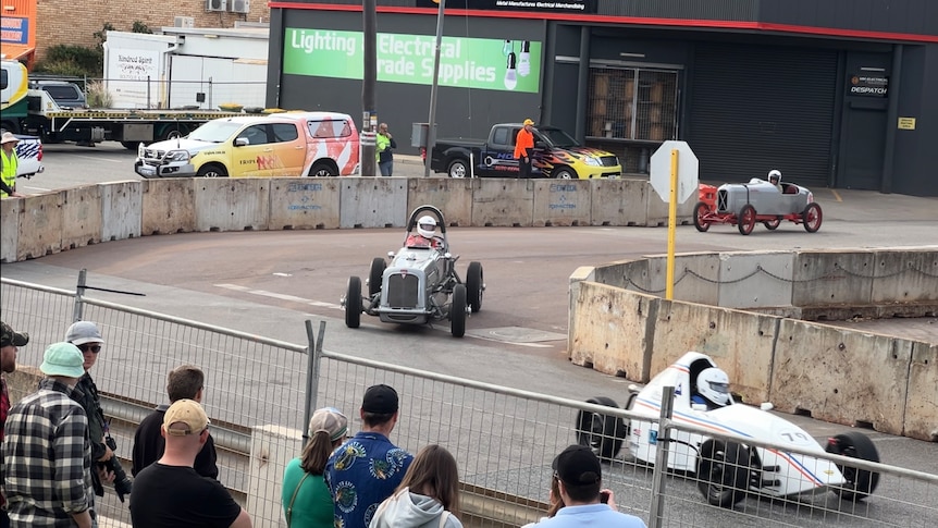 Three old style hot rods race on track surrounded by safety fencing and bollards