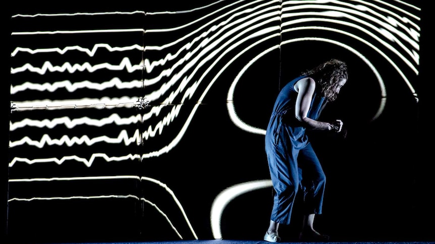 In front of a projection of a waveform, Jodee Mundy stands hunched over wearing a blue jumpsuit.