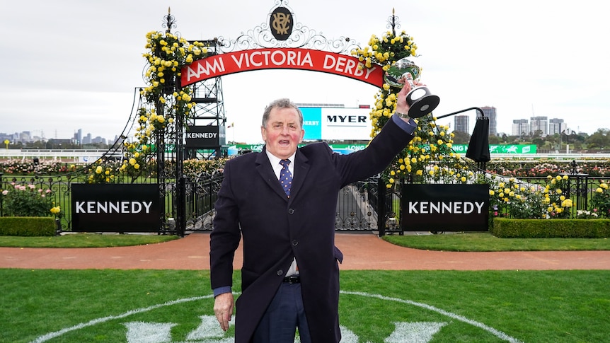 A man in a suit holds a trophy in his left hand as he stands in front of a sign at Flemington raceourse saying "Victoria Derby".