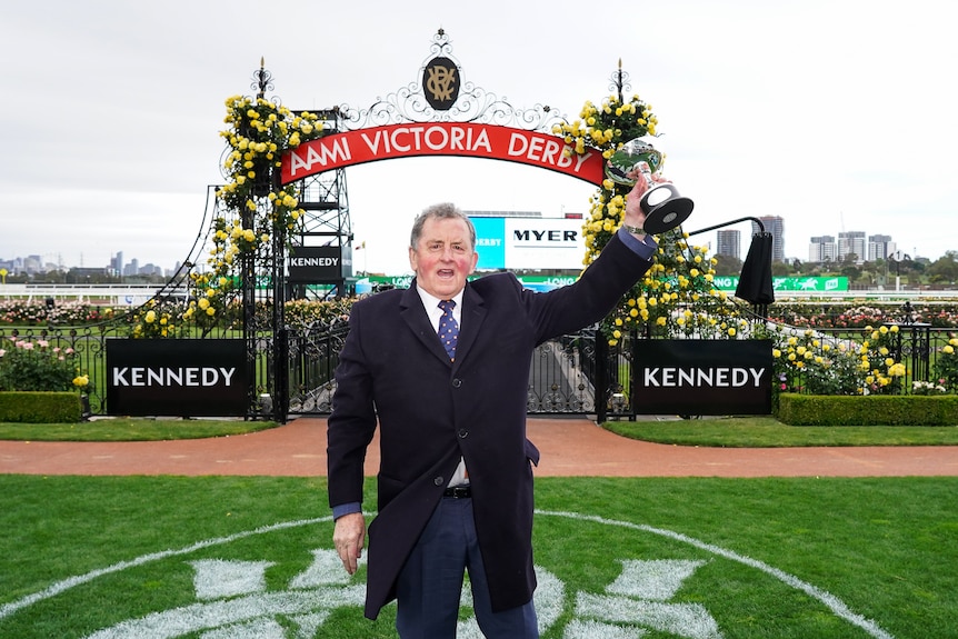A man in a suit holds a trophy in his left hand as he stands in front of a sign at Flemington raceourse saying "Victoria Derby".