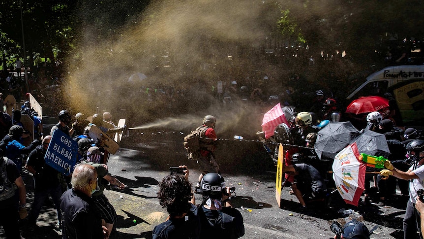 People are split on two sides some holding umbrellas and others holding signs as smoke and water fills the air