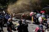 People are split on two sides some holding umbrellas and others holding signs as smoke and water fills the air