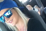 Kerri-Ann Conley with sunglasses and her hat backwards drives taking a selfie with a child sleeping in the car in the back.