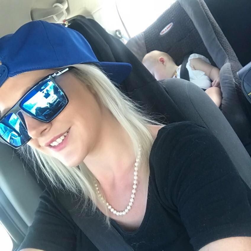 Kerri-Ann Conley with sunglasses and her hat backwards drives taking a selfie with a child sleeping in the car in the back.