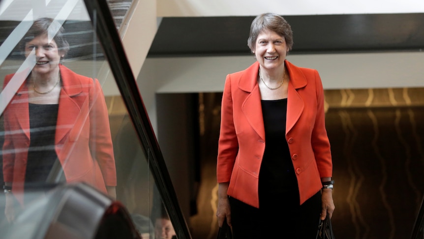 Helen Clark, wearing a red blazer over a black dress, smiles while standing on an escalator, her reflection in the glass