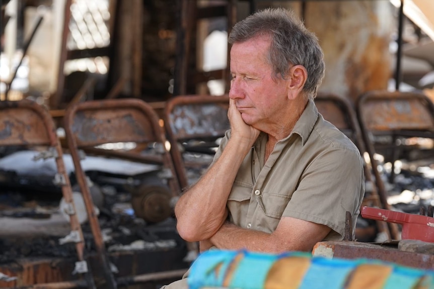 A man wearing a khaki shirt sits with his head on his hand with burnt metal chairs surrounding him.