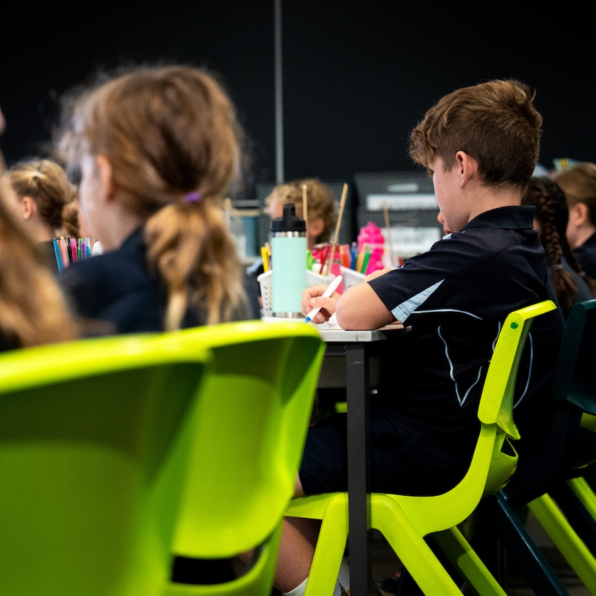 A row of students sit at their desks on green plastic chairs in a primary school classsroom.