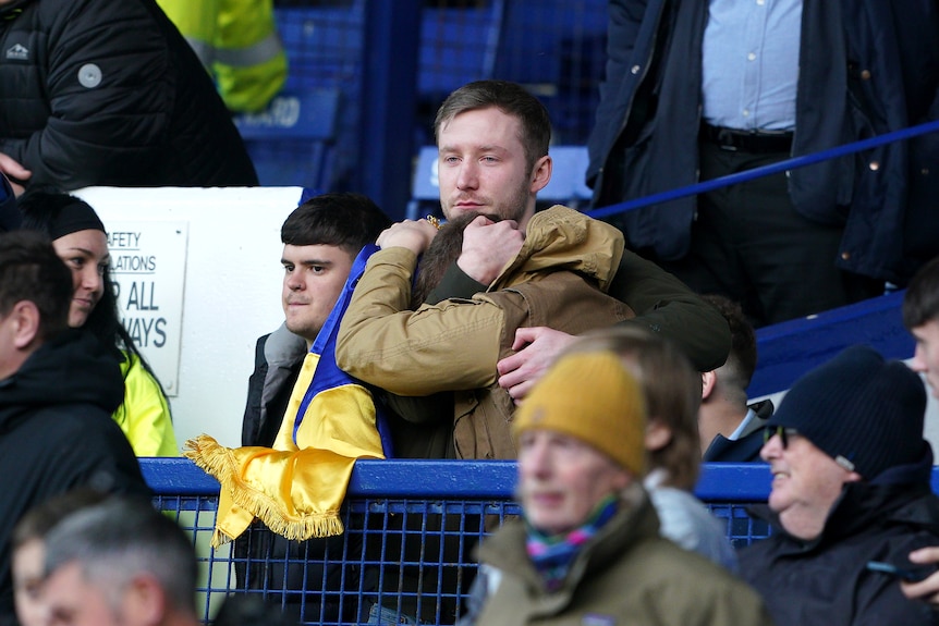 Emotional fans hug at a Premier League game Manchester City-Everton game. One holds a Ukraine flag.