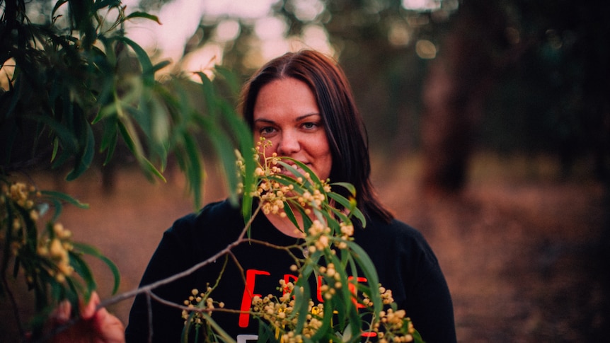 Rachael McPhail, who campaigned for Australia Post to include First Nations place names, smiling behind a wattle tree at sunset.