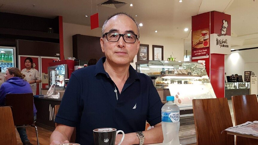 Medium close up of franchisee Wayne Hong sitting at a table at his Michel's Patisserie cafe, with coffee and water on table