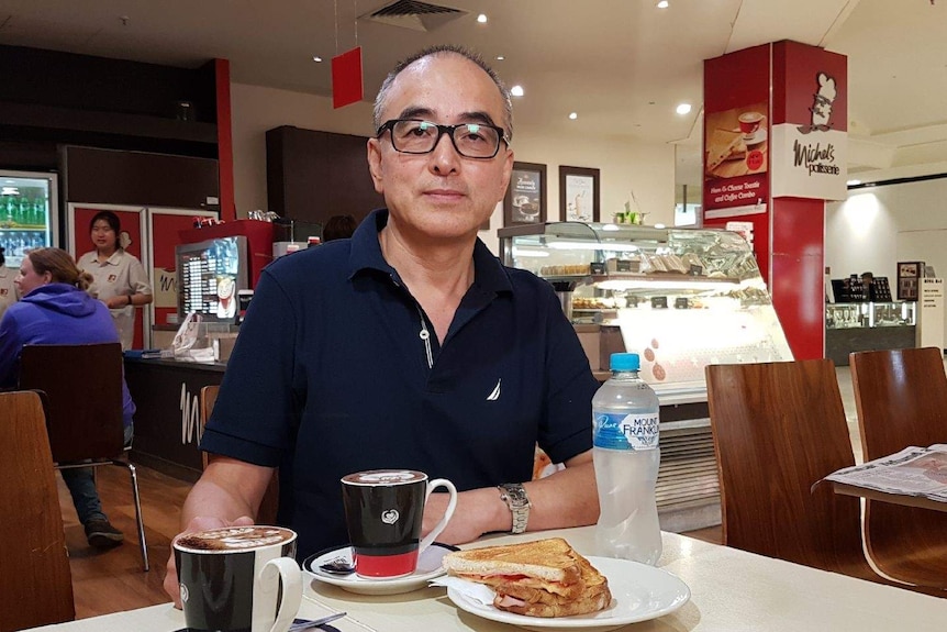 Medium close up of franchisee Wayne Hong sitting at a table at his Michel's Patisserie cafe, with coffee and water on table
