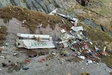 Aircraft remains and debris are scattered on the top of a rugged mountain with brown grass on the edges