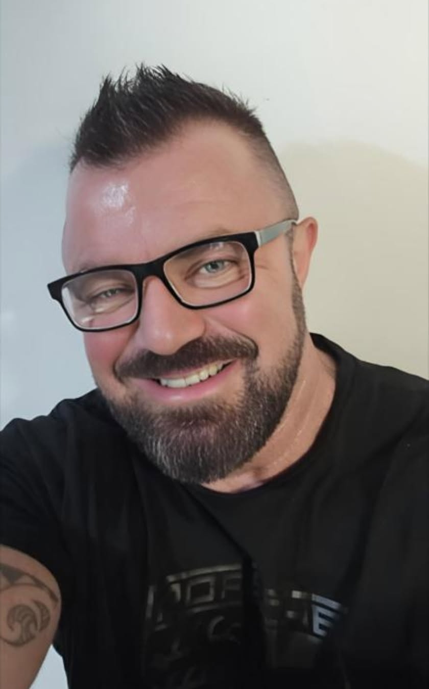 Uma selfie em close de George Napper usando óculos de armação preta e uma camiseta preta.