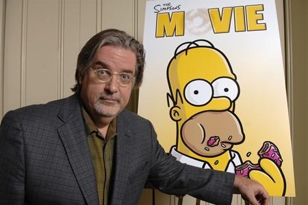 A bearded bespectacled man stands next to a poster for The Simpsons Movie.
