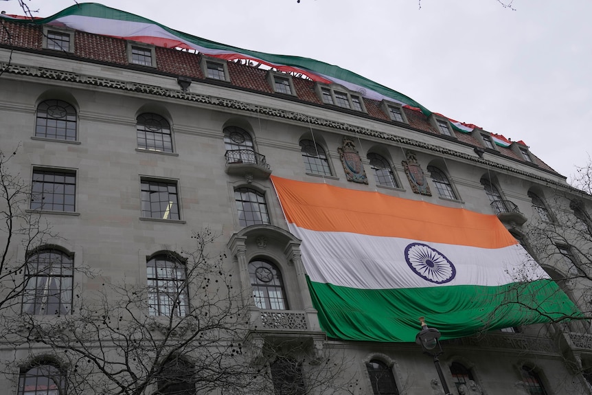 A two-storey-tall Indian flag hangs from balconies on a stone building, while people on its roof have another, longer flag.