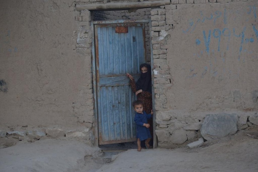 A woman and boy peak out from behind a blue door on a crumbling cement building.