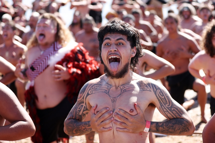 A warrior clutches both hands to his chest, while his mouth is wide open, during a haka