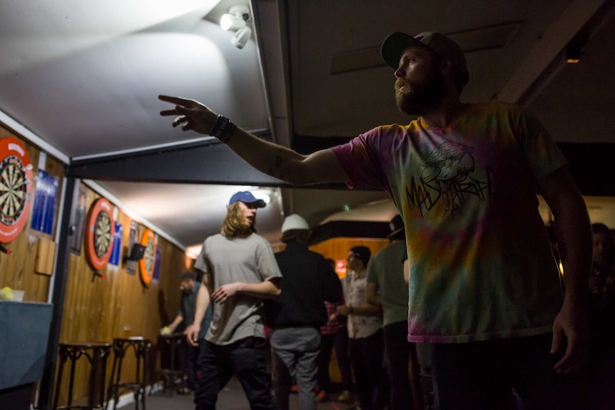 A bearded, capped staffer throws a dart as others socialise in the background.