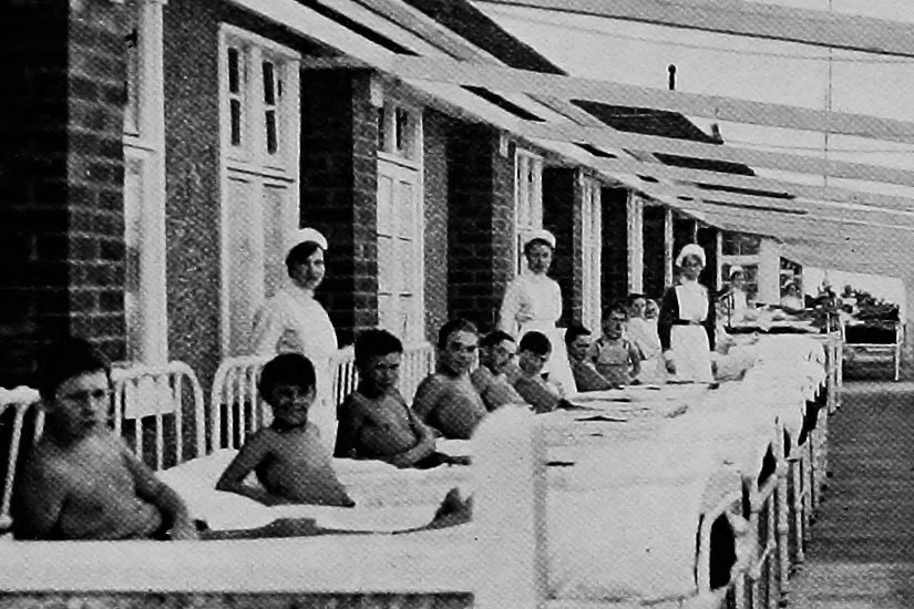 A black and white image of young boys lying in hospital beds on a long outdoor veranda