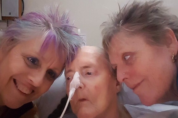 Two older women cuddle up to their elderly mother, who appears to be in an aged care home.