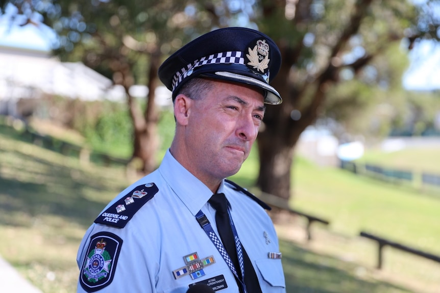  Police man wearing navy blue police hat speaks while standing in front of green sports field near trees