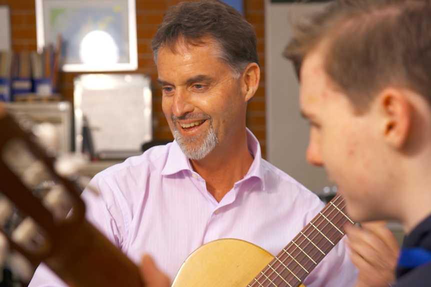 A man smiles as he and a boy sit to play classical guitar.
