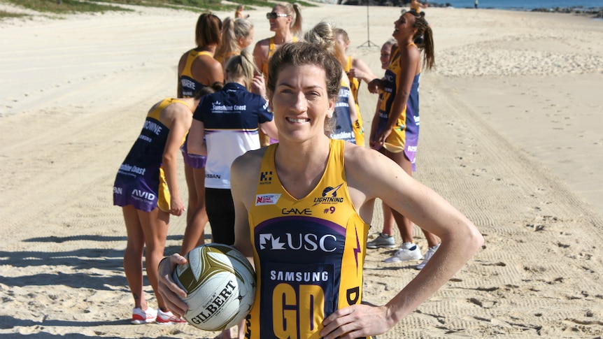 One netballer stands with a ball smiling on the beach, while teammates are behind talking, happy and laughing.