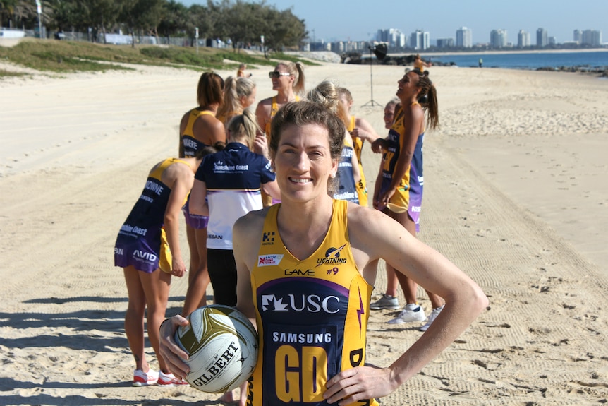 One netballer stands with a ball smiling on the beach, while teammates are behind talking, happy and laughing.