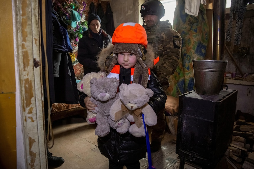 A girl is holding stuffed teddy bears wearing a bright orange helment and vest. 