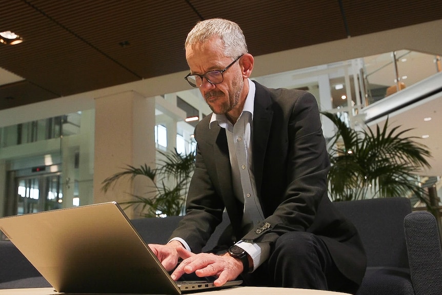 Man with cropped grey hair, stubble, glasses, black suit, open collar light shirt sitting at a table typing on a computer.