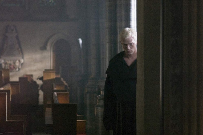A pale man with pure white hair wearing a brown monk's robe stands menacingly in a church corner.