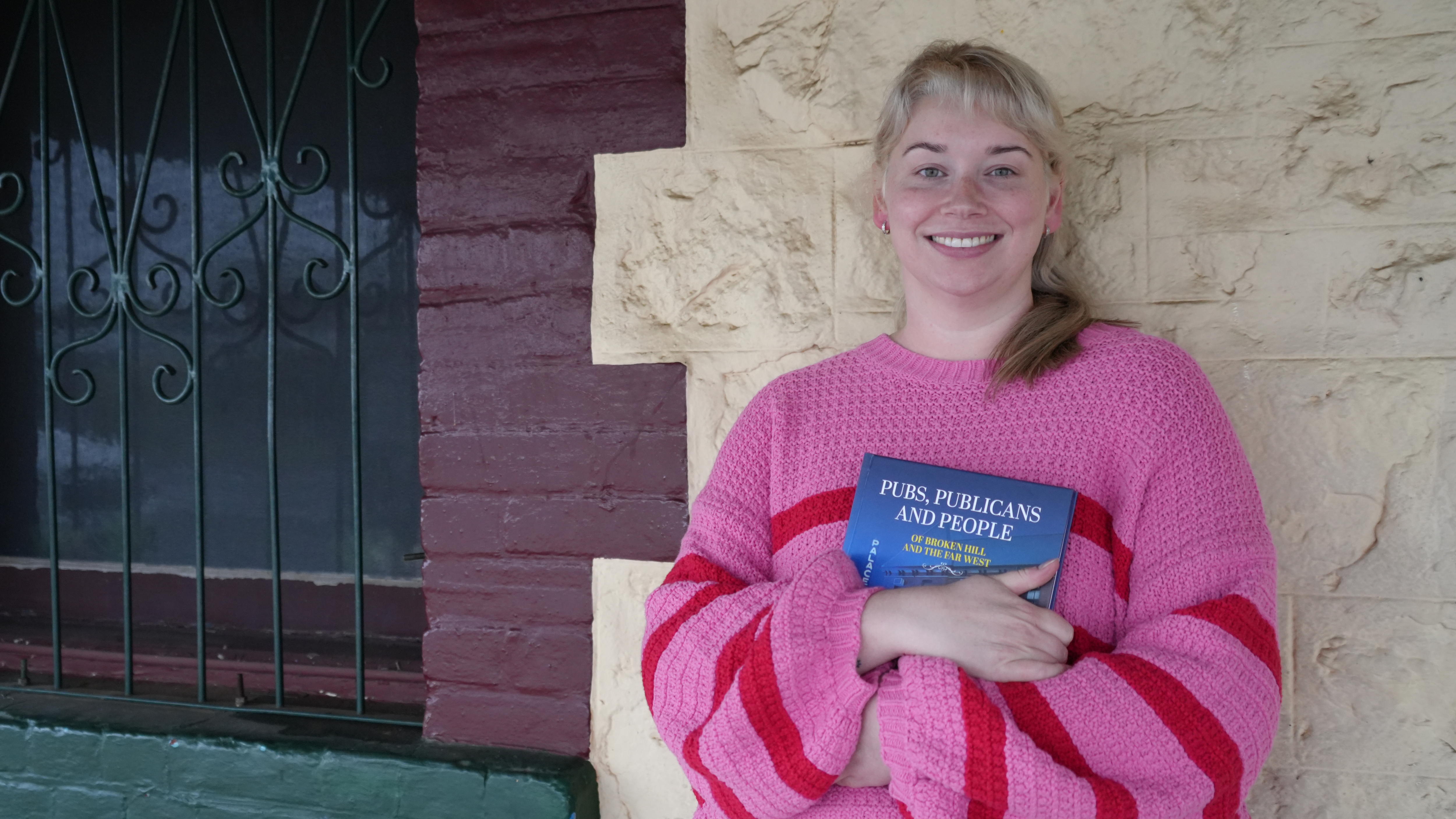 A blonde woman standing in front of a brick wall, wearing a colourful jumper and holding a book while smiling at the camera 