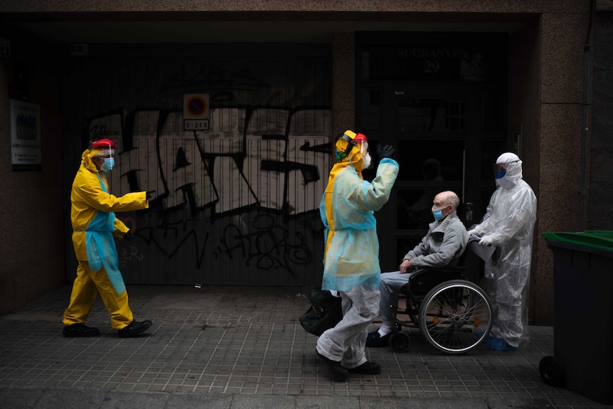 A person in protective clothing pushes an old man in a wheelchair wearing a mask.