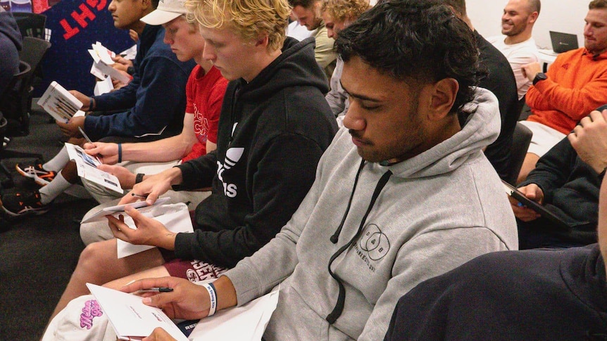 A pacific island rugby player looks at a form while sitting in a room with other players 