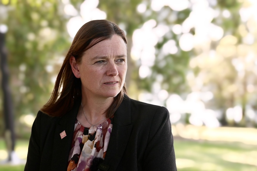 A woman with long, straight auburn hair, wearing a black jacket over a patterned shirt, is in parkland, looking to her left