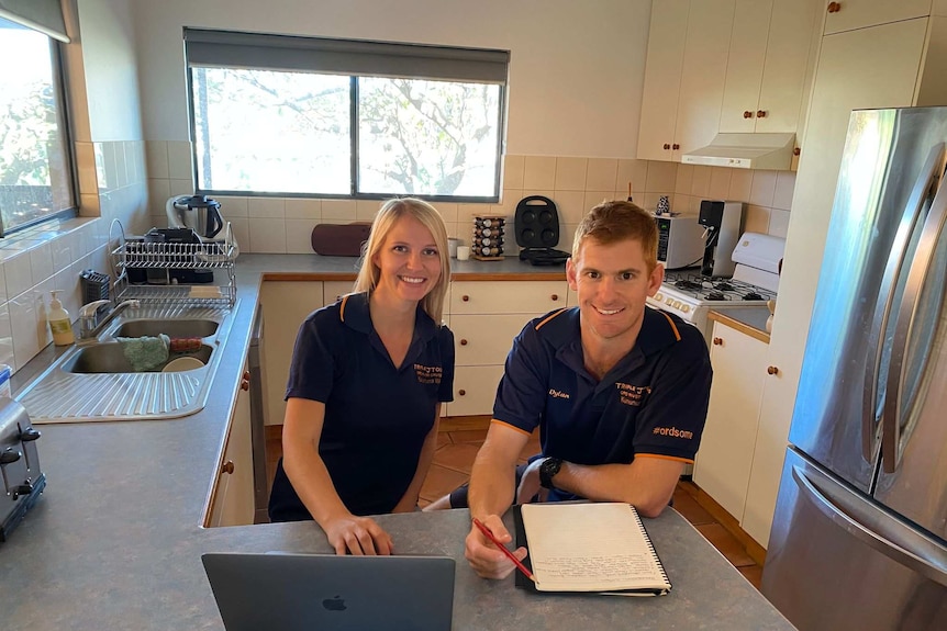 Chelsea and Dylan sitting at a kitchen bench wearing matching dark blue company polo shirts, in front of a laptop.