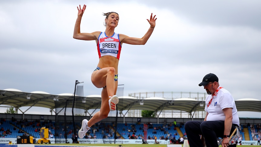 A female long jumper is pictured in mid jump with arms out and legs at full stretch above the sand. 