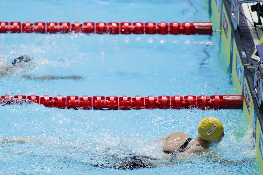 Ariarne Titmus, wearing a yellow swimming cap, touches the wall as Katie Ledecky swims behind.