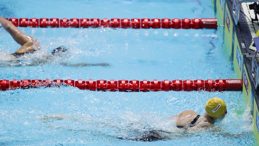 Ariarne Titmus, wearing a yellow swimming cap, touches the wall as Katie Ledecky swims behind.