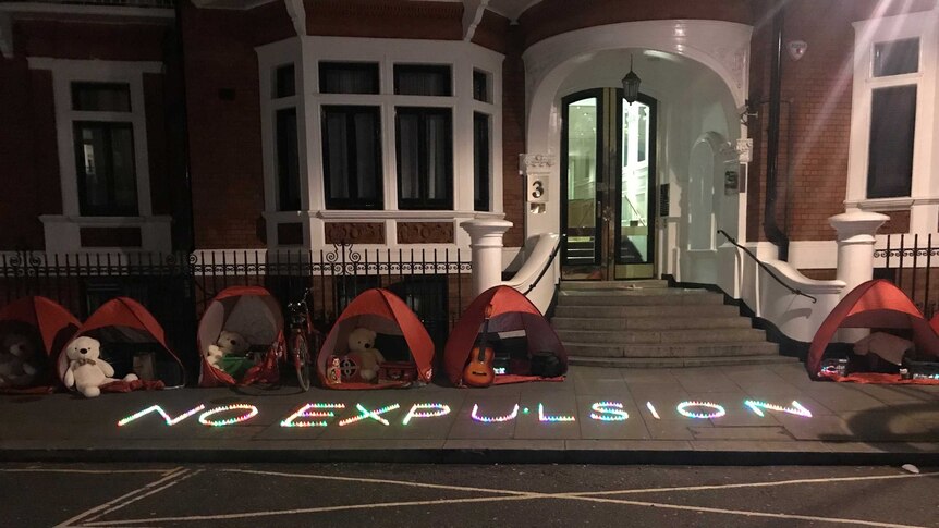 An illuminated sign on a pavement spells out 'No Expulsion' alongside protesters' tents outside an embassy