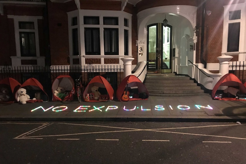An illuminated sign on a pavement spells out 'No Expulsion' alongside protesters' tents outside an embassy