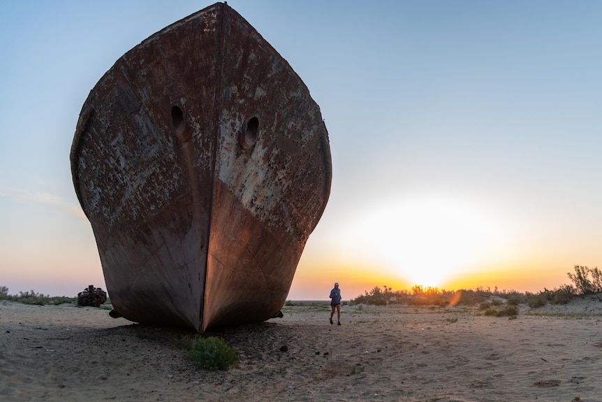 A woman runs next to a rusted ship that lies on what once was the bed of a sea, the sun sets behind. 