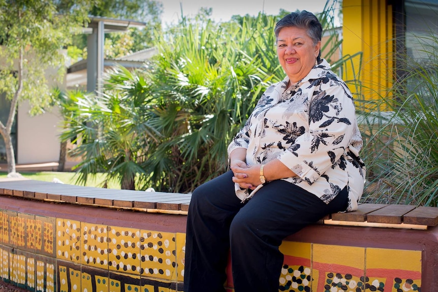 An Indigenous woman sits on a garden bench painted with traditional Aboriginal artwork amongst a native Australian garden.