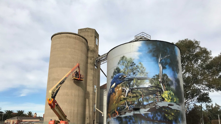 Two silos getting painted, the shorter of which is already decorated with an image of an azure kingfisher eating a golden perch